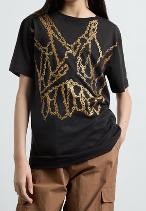 Person iført en sort T-shirt med mønster af metallic gyldne kæder og brune cargobukser, stående mod en ensfarvet lys baggrund.