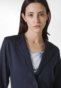 Marinblazer med slagkrage och enkel knappstängning, tillverkad av ett lätt material. Under blazer är det en ljusblå blus med blommönster.