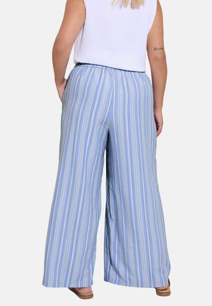 Person iført blå og hvide lodrette stribede bukser med vide ben og en ærmeløs hvid top, stående med hænderne i lommerne.