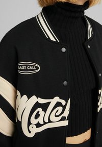 Schwarze Varsity-Jacke mit beigen Akzenten, geripptem Kragen und einem Front-Graphic "Last Call". Getragen über einem schwarzen gerippten Rollkragenoberteil.