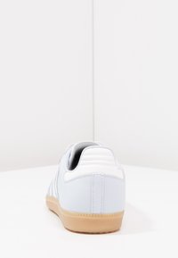 Světle modrá teniska s bílými doplňky, strukturovanou gumovou podrážkou a prošíváním na svršku. Pohled zezadu ukazuje polstrovaný límec paty.