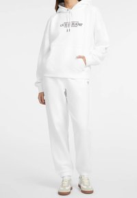 Sudadera con capucha blanca con bolsillo frontal, cordón y logo impreso. Combinada con pantalones jogger blancos y zapatillas claras con suela marrón.