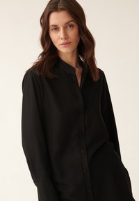 Schwarze langärmlige Bluse mit Knopfleiste und Stehkragen. Glatte Textur, lockere Passform und dezentem Glanz.