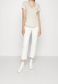 T-shirt beige a V, maniche corte, indossata con jeans bianchi a gamba dritta e sneakers bianche. Design semplice, look casual.