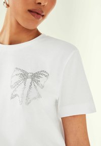 T-shirt en coton blanc avec un design de nœud en strass argentés sur le devant, à manches courtes et à encolure ronde classique.