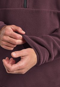 Burgundy fleece pullover with quarter-length zipper, featuring textured fabric and elasticized cuffs; hands adjusting sleeve length.