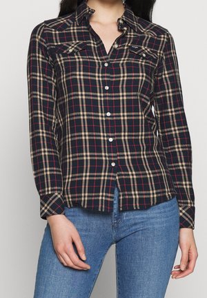Kvinde iført en sort, beige og rød ternet skjorte med knapper og to brystlommer, kombineret med blå jeans.