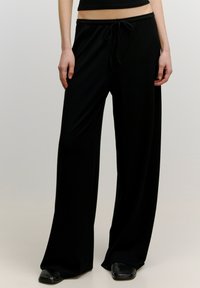 Zwarte wijde broek in ribbelstof met een trekkoord in de taille, gladde textuur, volle lengte en subtiele val. Gecombineerd met platte schoenen.