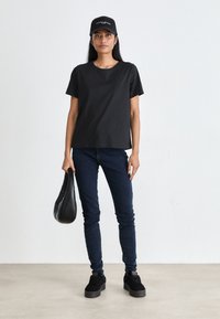 T-shirt noir à manches courtes, skinny jeans bleu foncé, baskets noires, tenant un sac noir, portant une casquette noire avec un texte blanc.