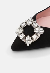 Chaussure en daim noir à bout pointu ornée d'une grande broche décorative en strass avec des accents de cristaux rectangulaires et ronds. Doublure intérieure rose pâle.