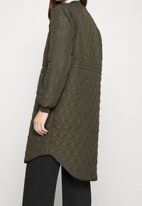 Cappotto lungo trapuntato verde oliva con un motivo a diamante, caratterizzato da un orlo leggermente arrotondato e polsini a costine, abbinato a pantaloni neri.