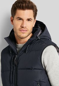 Marineblauer gefütterter Weste mit Kapuze, ausgestattet mit einem Reißverschluss, verstellbaren Kordeln und einer leichten, gesteppten Textur. Trägt über einem grauen Pullover.