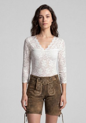 Vrouw met golvend donker haar, gekleed in een witte bloemenkanten top met driekwart mouwen en bruine traditionele geborduurde leren korte broek.
