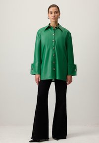 Zaļš mākslīgās ādas pogājams krekls ar norāvu plati apmali, garām piedurknēm un platām aprocēm, kas rotātas ar dekoratīvām pogām. Apģērbs apvienots ar melnām platām biksēm.