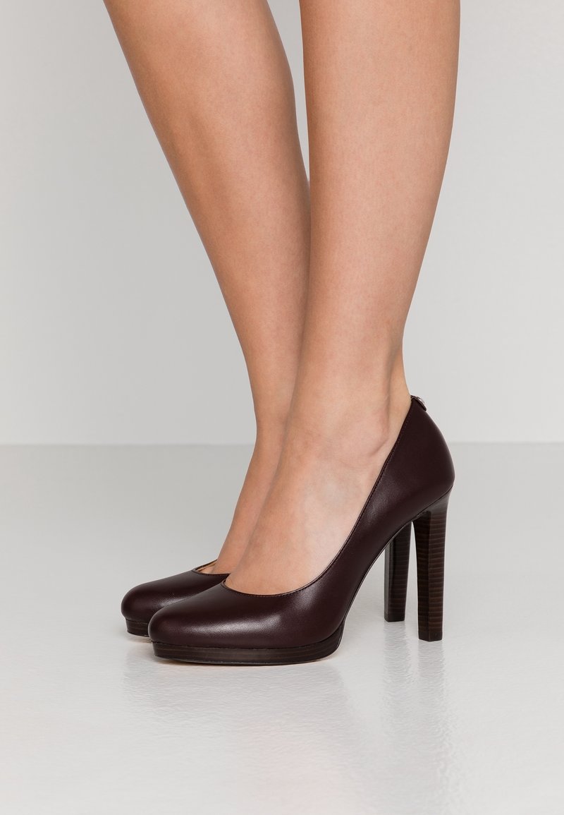 Femme portant des escarpins en cuir marron foncé à talons hauts, debout sur un sol clair et brillant, jambes visibles du milieu du mollet vers le bas.
