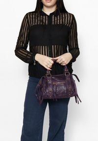 Paarse leren handtas met metallic accenten, franjes en een gestructureerd ontwerp. Draagdoor een persoon in een zwart gestreepte blouse en blauwe broek.