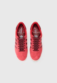 Red suede sneakers featuring three maroon stripes, round toe, and a textured white rubber sole. Inside has a red pattern with branding.