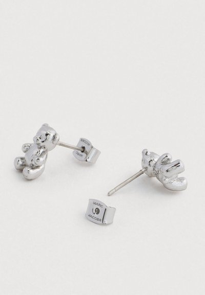 Stříbrné náušnice ve tvaru medvěda s lesklým povrchem, s uzávěrem na tyčince a značkou "Marc Jacobs" na zadní straně.