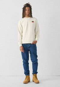 Sudadera crema con un pequeño logo rojo y negro, combinada con jeans azules y botas marrones. El diseño es casual con puños y dobladillo acanalados.