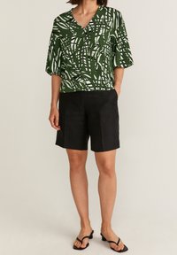 Blusa a maniche corte verde e bianca a fantasia con scollatura a V, abbinata a pantaloni corti neri su misura e sandali flip-flop neri.