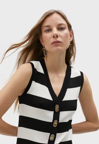 Czarny i biały, paskowany, bezrękawnik z dzianiny z dekoltem w kształcie litery V. Posiada pięć metalowych guzików z przodu oraz gładką teksturę.