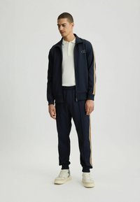 Marineblauer Trainingsanzug mit cremefarbenen Streifen, bestehend aus einer Reißverschlussjacke und Jogginghosen mit elastischem Bund, kombiniert mit beigen Sneakern und einem weißen Poloshirt.
