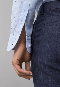 Main droite à côté de la poche d'un pantalon bleu foncé texturé, portant une chemise bleu clair avec un petit motif sombre et un poignet boutonné.
