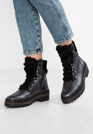 Ben i opsmøgede blå jeans iført sorte teksturerede læder snørestøvler med tykke såler og sort pelyderfoering på hvidt gulv.