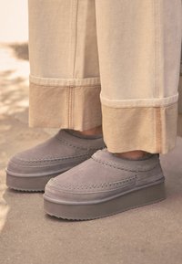 Grå ruskinds slip-on sko med platformsål, der har dekorativ syning og en blød, struktureret overflade, parret med beige vidde ben bukser.