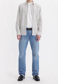 T-shirt bianco indossato sotto una camicia a righe beige e grigia, abbinato a jeans a gamba dritta blu chiaro e mocassini neri.