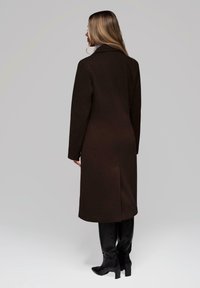 Mujer de pie con la espalda hacia la cámara, vistiendo un abrigo largo marrón oscuro y botas negras hasta la rodilla, contra un fondo liso.