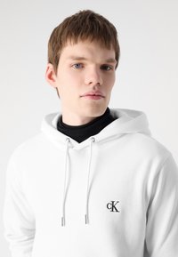 Sudadera blanca de tejido suave con capucha ajustable y un pequeño logotipo negro bordado en el lado izquierdo del pecho.