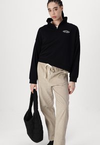 Sudadera negra con cremallera y un logo, combinada con pantalones de pana beige claro y un bolso de mano negro acolchado. Tejido suave y corte relajado.