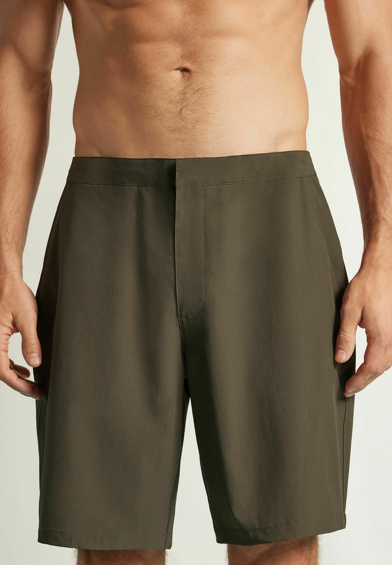 Buste masculin portant un short vert olive jusqu’aux genoux, debout, bras détendus le long du corps, sur un fond clair uni.