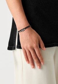 Svart flettet armbånd med sølvdetaljer, som har en løkket design. Hånd plassert mot en svart topp og hvitt plissert stoff.