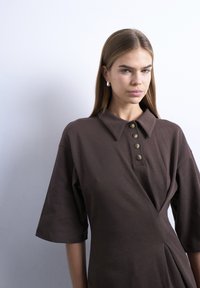 Mlada ženska s svetlo rjavimi ravnimi lasmi nosi temno rjavo obleko s ovratnikom in štirimi gumbi ter srebrne trikotne uhane.