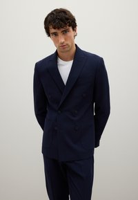 Blazer blu navy doppiopetto con rever a punta, due tasche frontali, sei bottoni e una texture liscia, abbinato a una maglietta bianca.