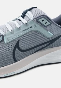La scarpa da corsa Nike Pegasus 40 presenta una tomaia in mesh grigia con un accento blu chiaro. Ha una suola imbottita e un logo swoosh di colore grigio scuro.