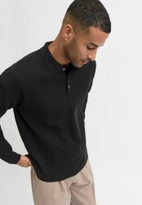 Fekete hosszúujjú pólót és bézs nadrágot viselő férfi, aki lefelé néz, egyik kezét a zsebében tartja, egy sima fehér háttér előtt.