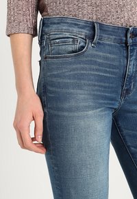 Vaqueros de mezclilla azul oscuro con un ligero desgaste, diseño de cinco bolsillos y un corte ajustado. Detalles cosidos a lo largo de los bolsillos y las costuras.
