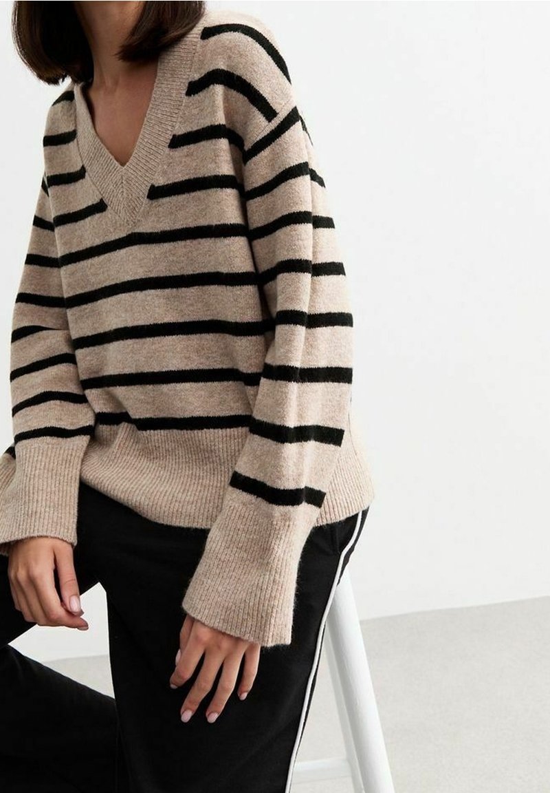 Pull rayé brun avec des rayures noires horizontales, design en V, manches longues et texture côtelée, associé à un pantalon noir avec des rayures sur les côtés.