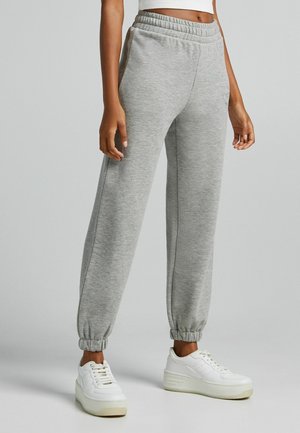 Personne portant un pantalon de jogging gris à taille élastique et des baskets blanches à plateforme, debout devant un fond gris uni.