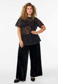 Top in pizzo nero con maniche corte sopra una blusa nera uniforme, abbinato a pantaloni larghi in velluto nero e scarpe slip-on nere.