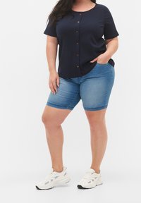 Camicia a righe blu navy con maniche corte e bottoni davanti, abbinata a shorts in denim azzurro. Il modello indossa sneakers bianche.