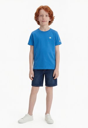 Dreng med rødt hår iført en blå Champion T-shirt, mørke denimshorts og hvide sneakers, stående mod en ensfarvet hvid baggrund.