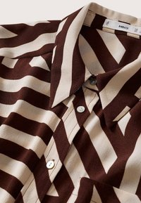 Chemise à col avec de larges rayures géométriques en zigzag marron et beige, boutons blancs sur le devant, étiquette en tissu visible à l'intérieur du col.