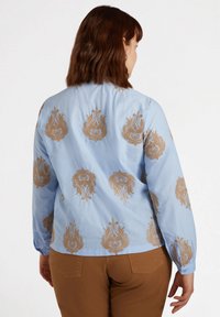 Hellbluse mit goldenen, gestickten Blumenmustern, langen Ärmeln und einem runden Saum. Glattes Material mit einem leichten Glanz.