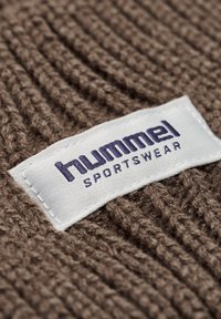 Bruine textuur stof met een wit label waarop "hummel" en "SPORTSWEAR" in paars staat. Gestikt voor duurzaamheid. Close-up weergave.