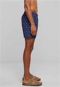 Shorts de bain navy avec des motifs nautiques blancs, taille élastique et cordon de serrage. Associés à des sandales en daim marron avec deux brides.