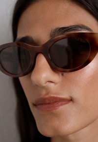Ochelari de soare tortoiseshell maro, cu o formă rotundă, lentile închise la culoare și un finisaj lucios, accentuând un design aerodinamic.